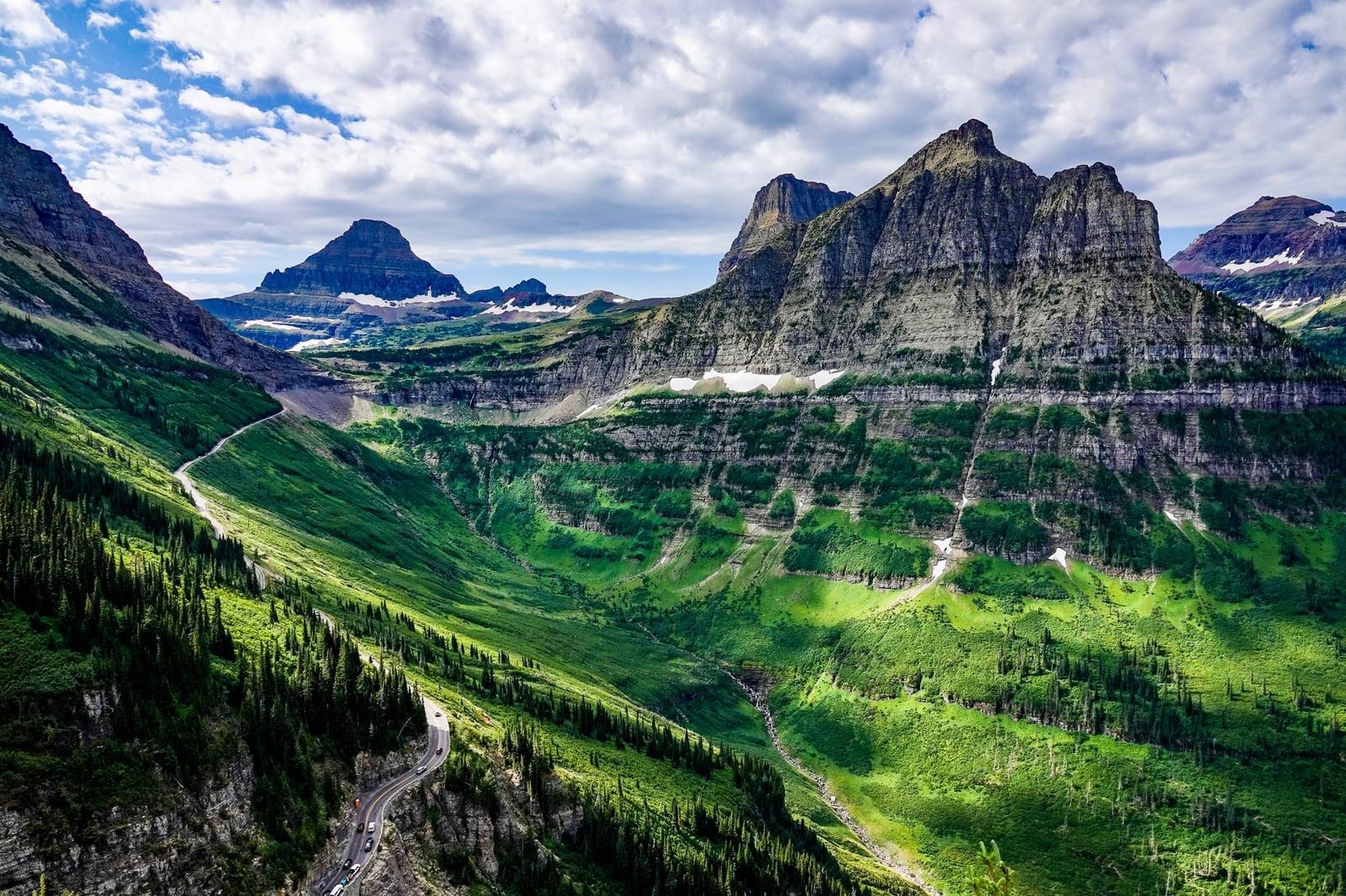 Going-to-the-Sun Road curves along steep green mountainsides in Glacier National Park beneath a partly cloudy sky.
